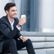 Attractive young businessman is sitting on steps outdoors. He is drinking coffee and smiling. The guy is looking aside with joy. Copy space in right side